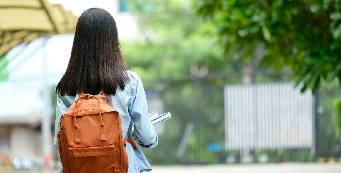 Girl standing infront of school gates with backpack and books in hand